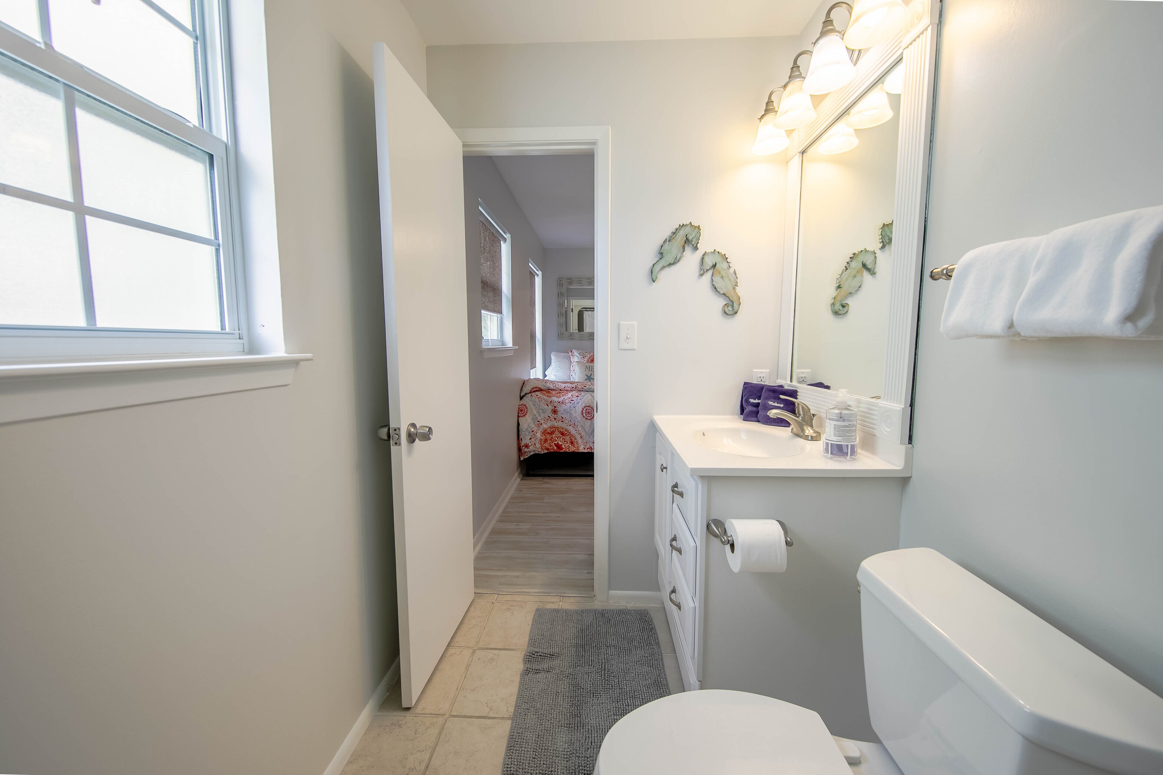 A bathroom with a toilet, sink, and mirror. There is a window and a door leading to another room.