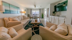A spacious living room with a large sofa, a coffee table, and a dining area with chairs and a table. There is a ceiling fan, a TV mounted on the wall, and a view of the ocean through the windows. - 30A Gulf front 2 Bed/2 Bath Sleeps 6 Private beach- Beach service included (Santa Rosa Beach)