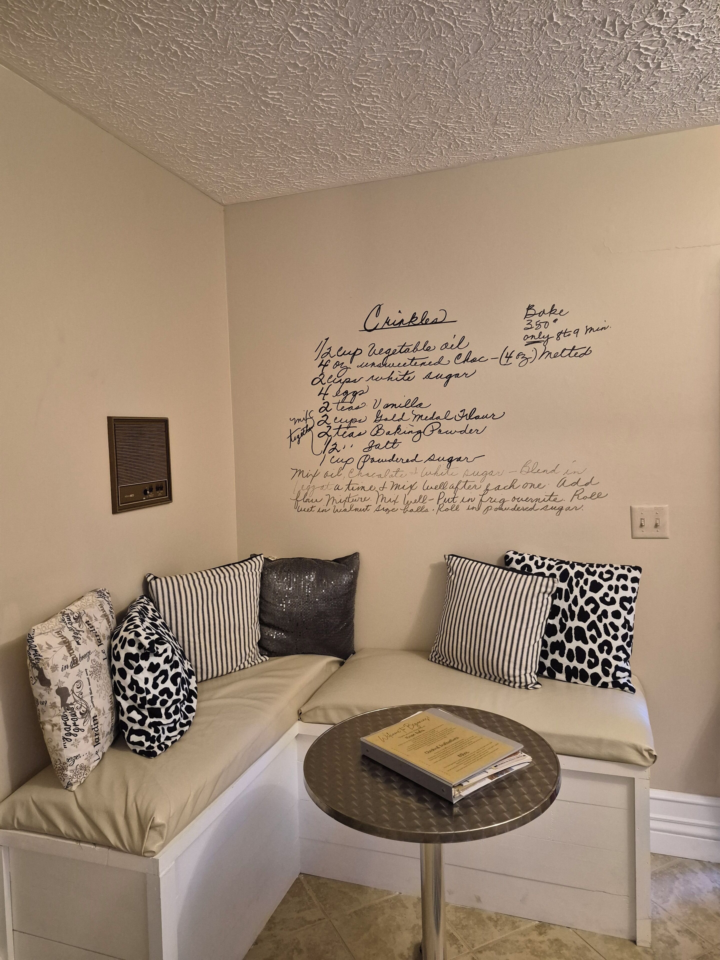 Un rincón acogedor con un espacio de asientos, una receta de 'crinkles' colgada en la pared y una pequeña mesa redonda con un libro.