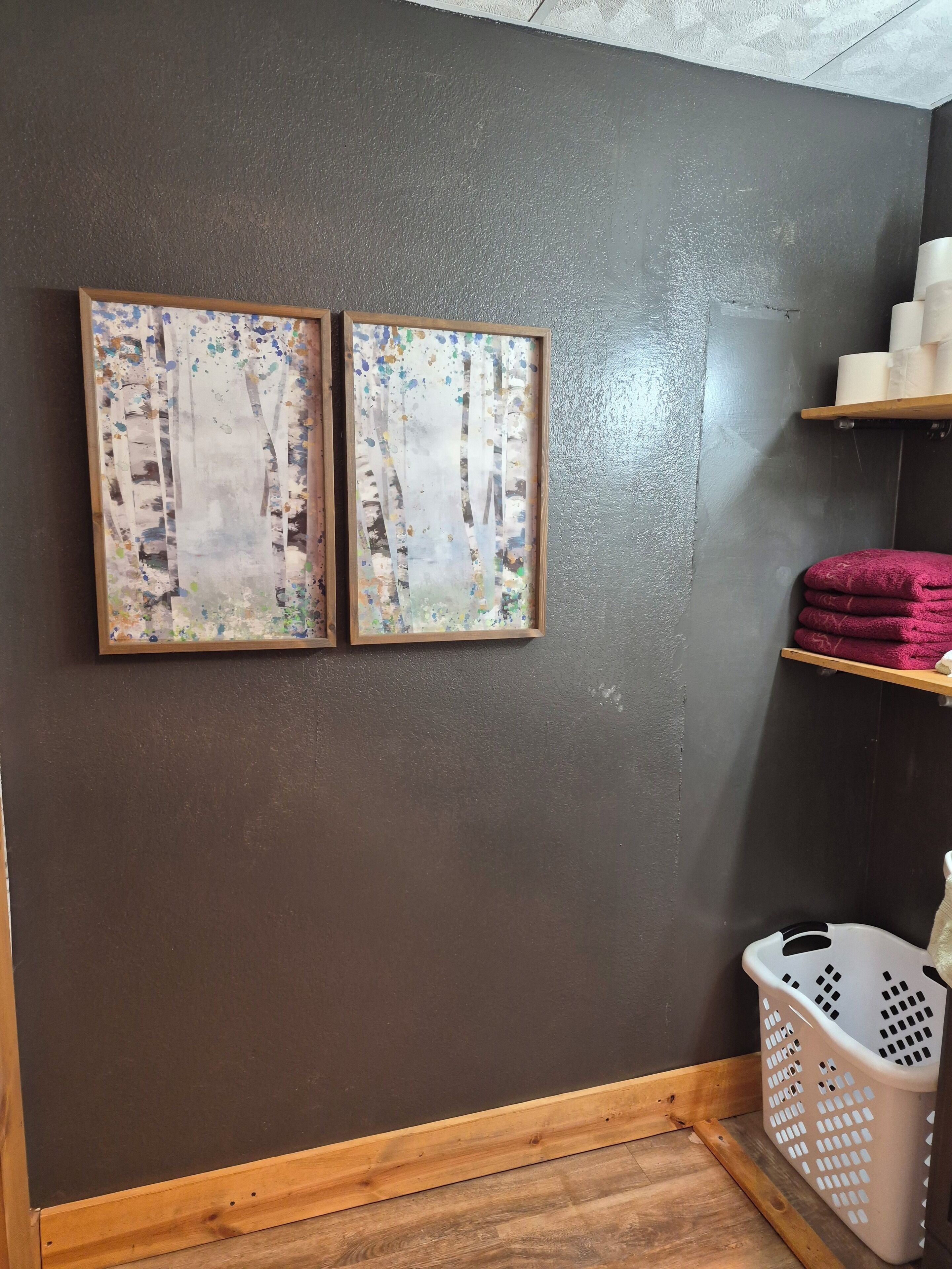 A room with a dark wall, two framed artworks, a wooden floor, a laundry basket, and shelves with towels.