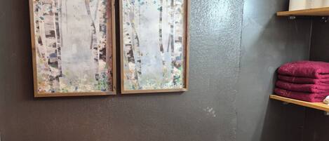 A room with a dark wall, two framed artworks, a wooden floor, a laundry basket, and shelves with towels.