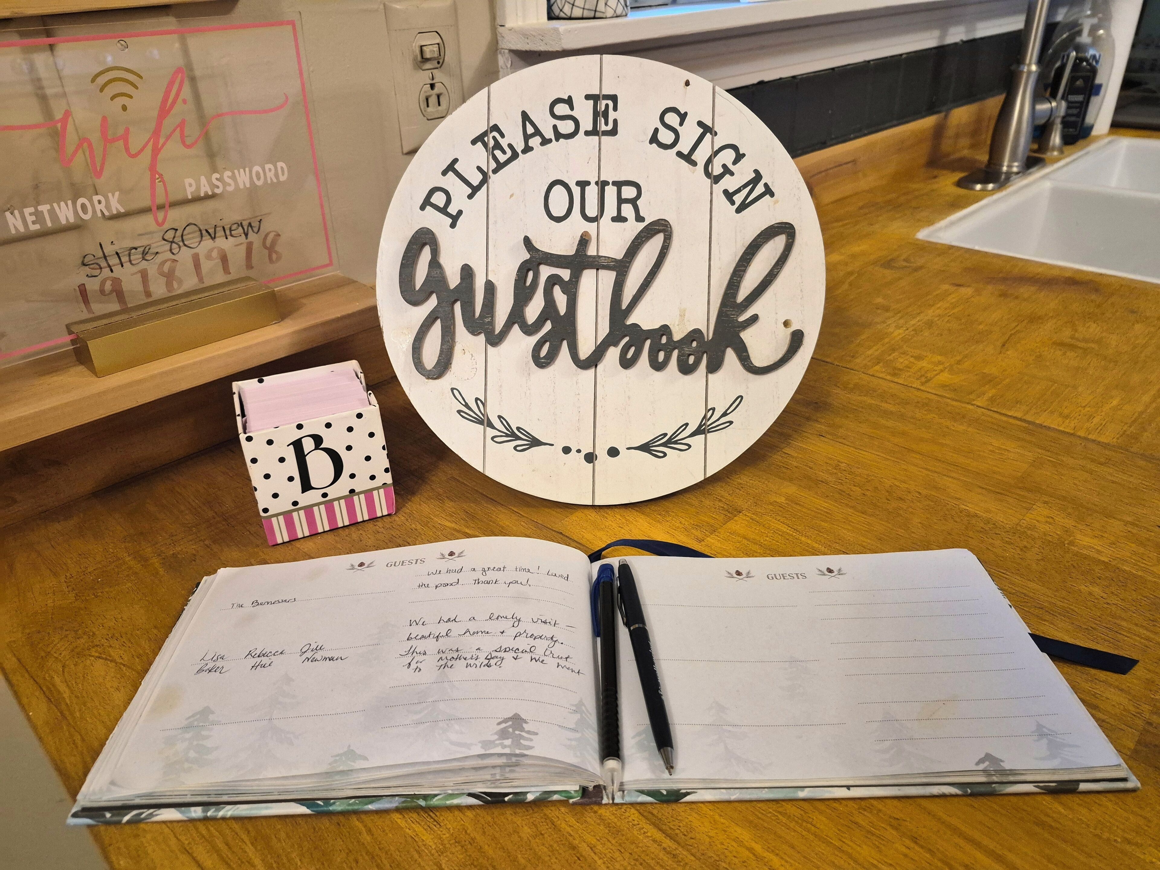Een rond bord met de tekst 'PLEASE SIGN OUR guestbook', een notitieboekje met handgeschreven aantekeningen en een klein doosje met de letter 'B' op een houten tafel.