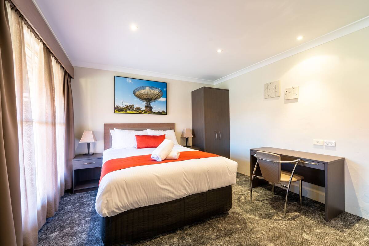 A hotel room with a bed, desk, chair, nightstand, lamp, and artwork on the wall.
