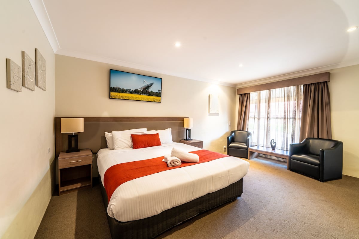 A hotel room with a large bed, a desk, a chair, a painting, and a window with curtains.