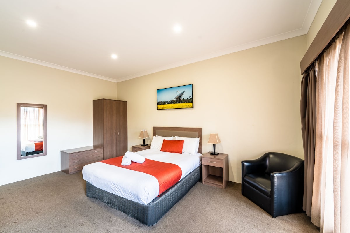 A hotel room with a large bed, a wardrobe, a desk with a lamp, a chair, and a painting on the wall.