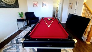 Ein Billardtisch in einem Raum mit modernem Design, inklusive eines schwarzen Stuhls, eines kleinen Tisches mit Pflanze und gerahmten Schmetterlingsbildern an der Wand.