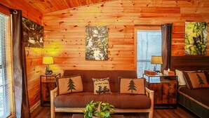 A cozy cabin interior with a wooden wall, a sofa with forest-themed cushions, a coffee table with a plant, and a bed with a forest mural. - Bigfoot Lodge Room One (Benton)