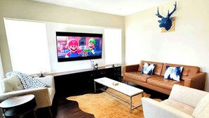 A living room with a flat-screen TV, a brown leather sofa, a white armchair, a small round table, and a cowhide rug. - Cozy Home near Alamodome~Free Gated Parking~Kids Climbing Dome & Backyard games (San Antonio)