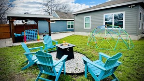 A backyard with a fire pit, four Adirondack chairs, a play structure, and a covered patio area. - Cozy Home near Alamodome~Free Gated Parking~Kids Climbing Dome & Backyard games (San Antonio)