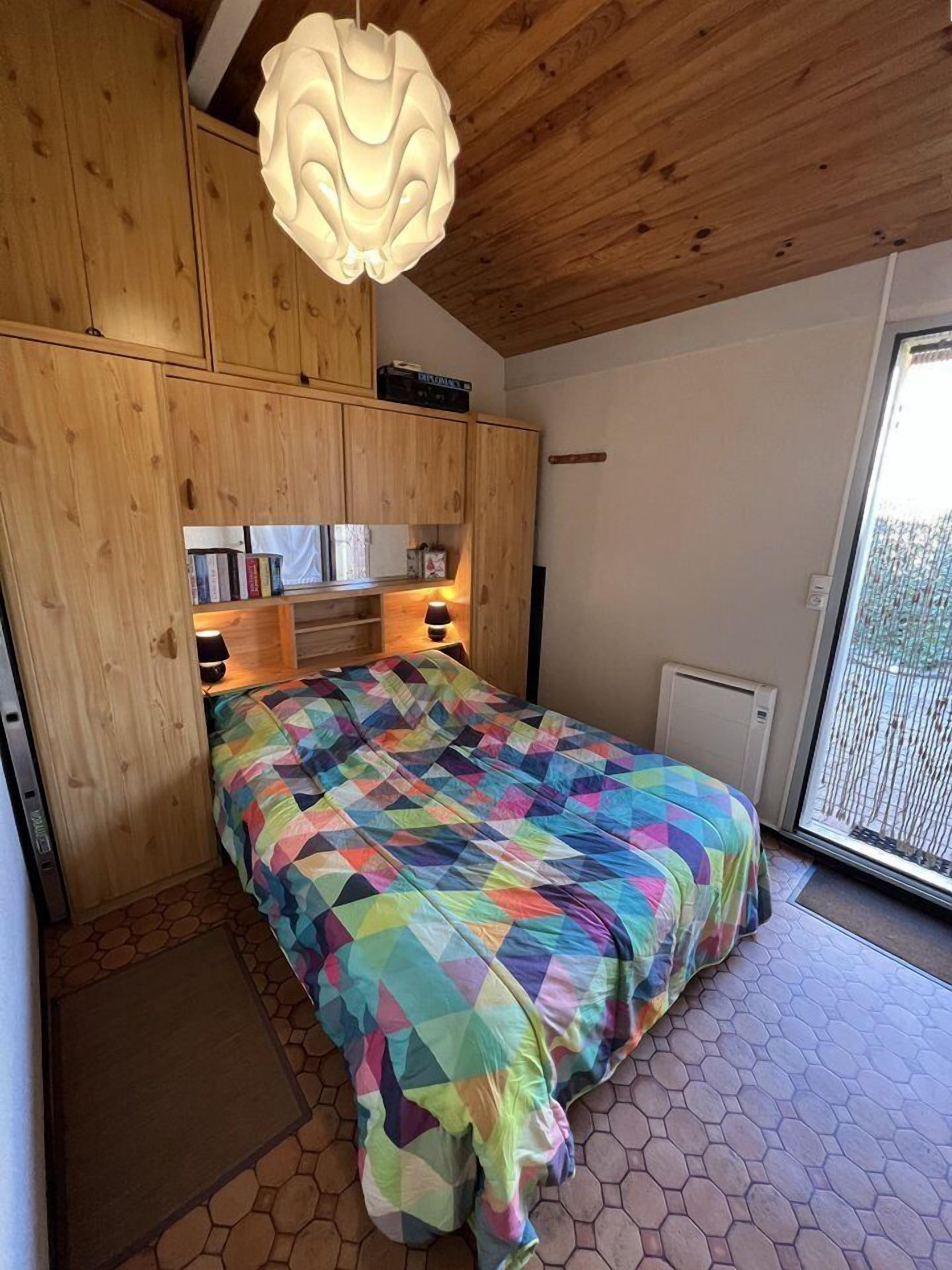 Een slaapkamer met een kleurrijke, geometrische sprei, houten kasten, een plafondlamp en een uitzicht door een glazen deur.