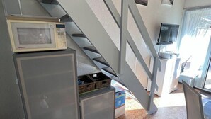 Uma cozinha compacta com micro-ondas, frigorífico e um armário pequeno. Uma escadaria leva a uma varanda com televisão.