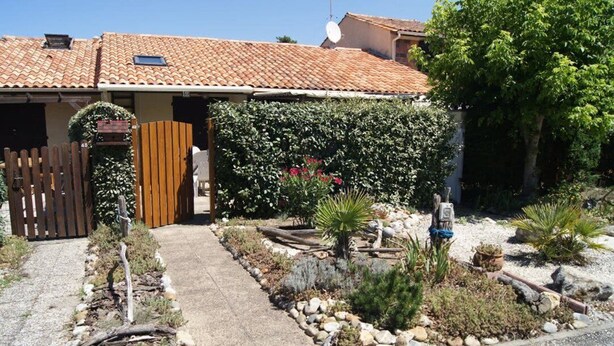 Uma casa com telhado de telhas, portão de madeira e um jardim com várias plantas e pedras.