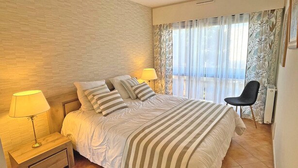 Ein Hotelzimmer mit einem Bett, zwei Nachttischlampen, einem Sessel und einem Fenster mit durchsichtigen Vorhängen.