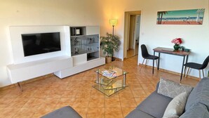 Ein Wohnzimmer mit Flachbildfernseher, einem Glastisch, einer Couch, einem Esstisch mit Stühlen und einer Topfpflanze.