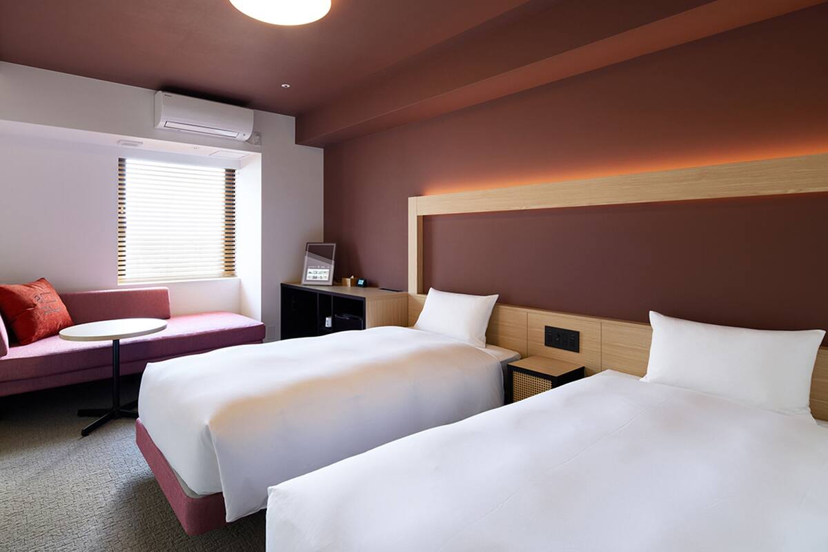 Une chambre d’hôtel avec deux lits, un petit canapé, une table et une fenêtre avec des stores.