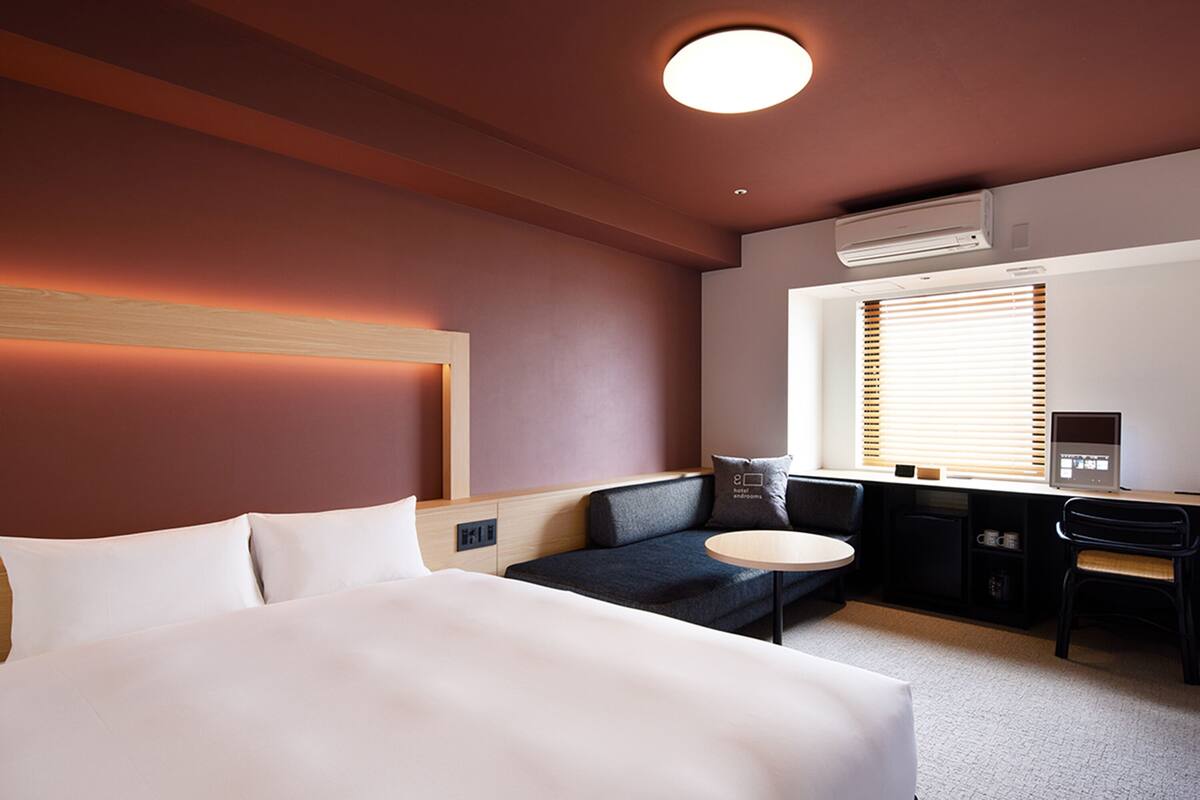 Une chambre d’hôtel avec un grand lit, un bureau, une chaise, une petite table ronde et une fenêtre avec des stores.
