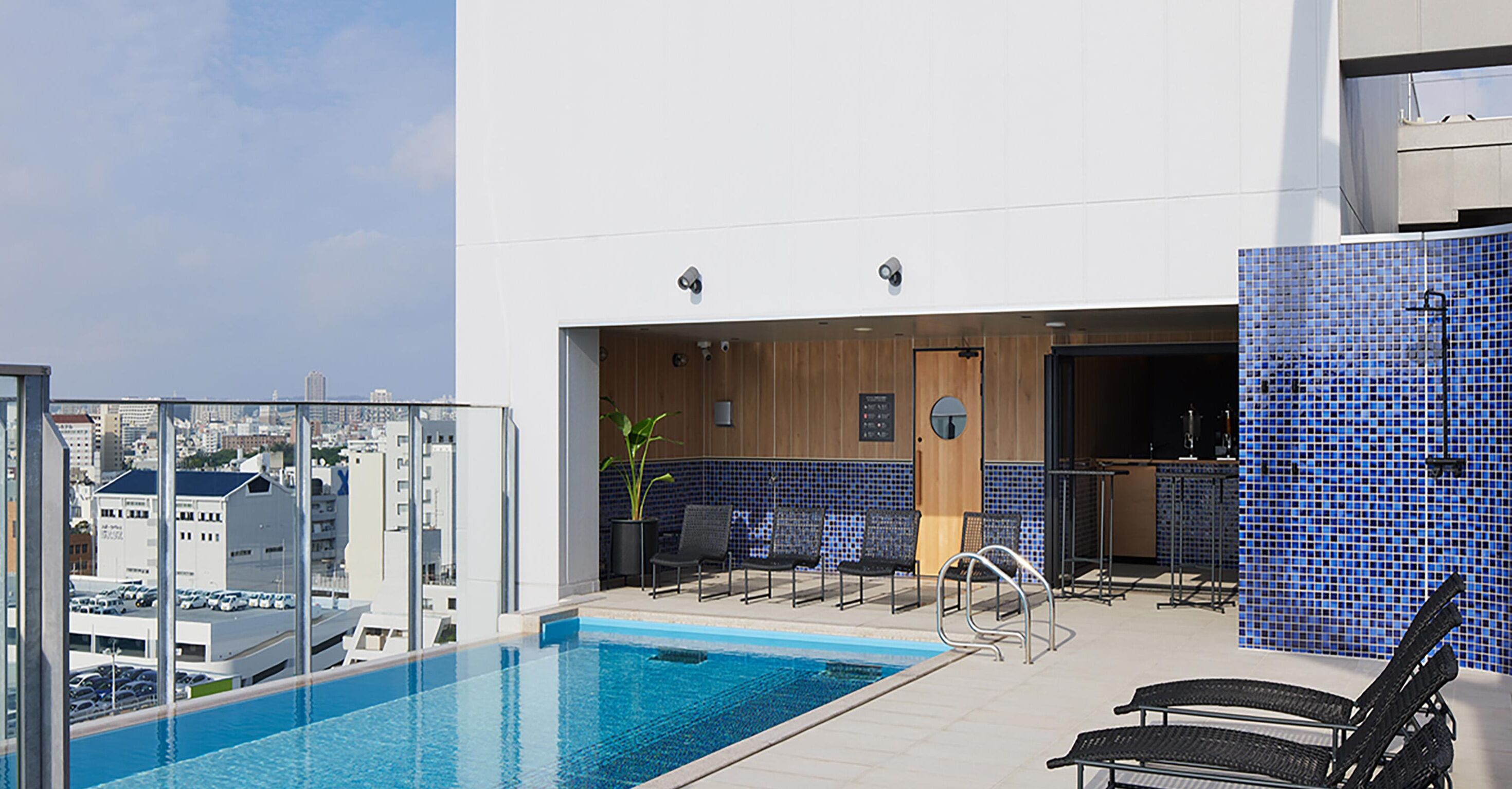 Un espace piscine aménagé sur le toit, offrant une vue dégagée sur le paysage urbain, un bar avec des sièges, et une porte en bois donnant accès à un espace intérieur.