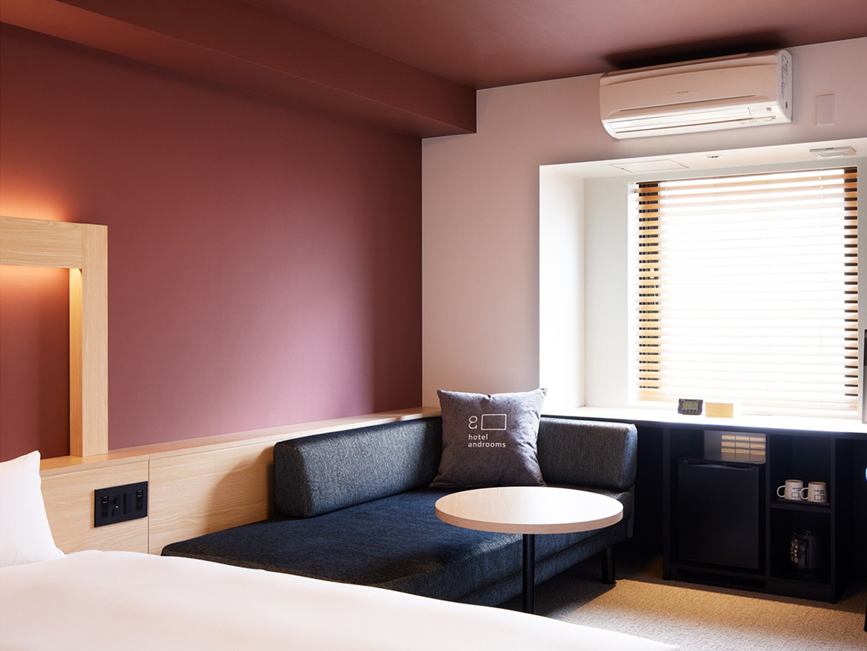 Una habitación de hotel moderna con cama, sofá, una mesita redonda y ventana con persianas.