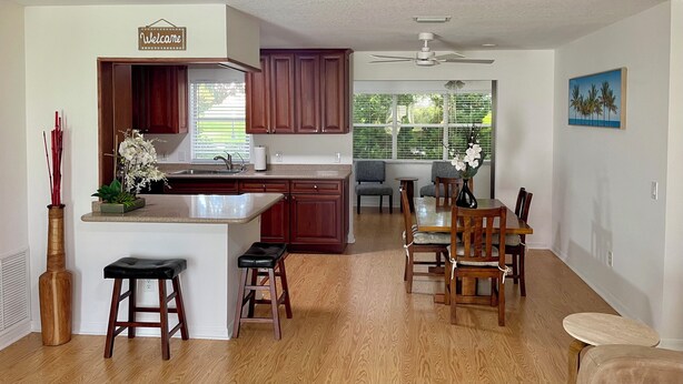 A modern kitchen with wooden cabinets, a central island, bar stools, and a dining area with chairs. - Private heated in-ground pool oasis with golf cart in Rio Grande! (The Villages)