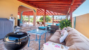 A covered outdoor patio with a grill, seating area, and a view of a swimming pool.