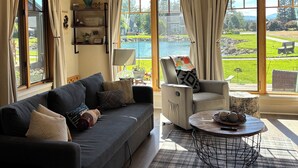 Un salon comprenant un canapé, un fauteuil et une table basse, de grandes fenêtres équipées de stores, un ventilateur de plafond et une vue sur un paysage verdoyant.