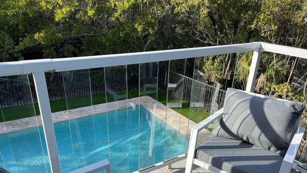 A poolside area with a glass-enclosed pool, a white deck, and a cushioned outdoor seating area. - New* Nature view #2/ 1 BED pool, walk to beach (Fort Myers Beach)