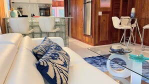 A modern living room with a white sofa, blue patterned pillows, a glass coffee table, and a dining area with white chairs and a glass table. - New* Nature view #2/ 1 BED pool, walk to beach (Fort Myers Beach)