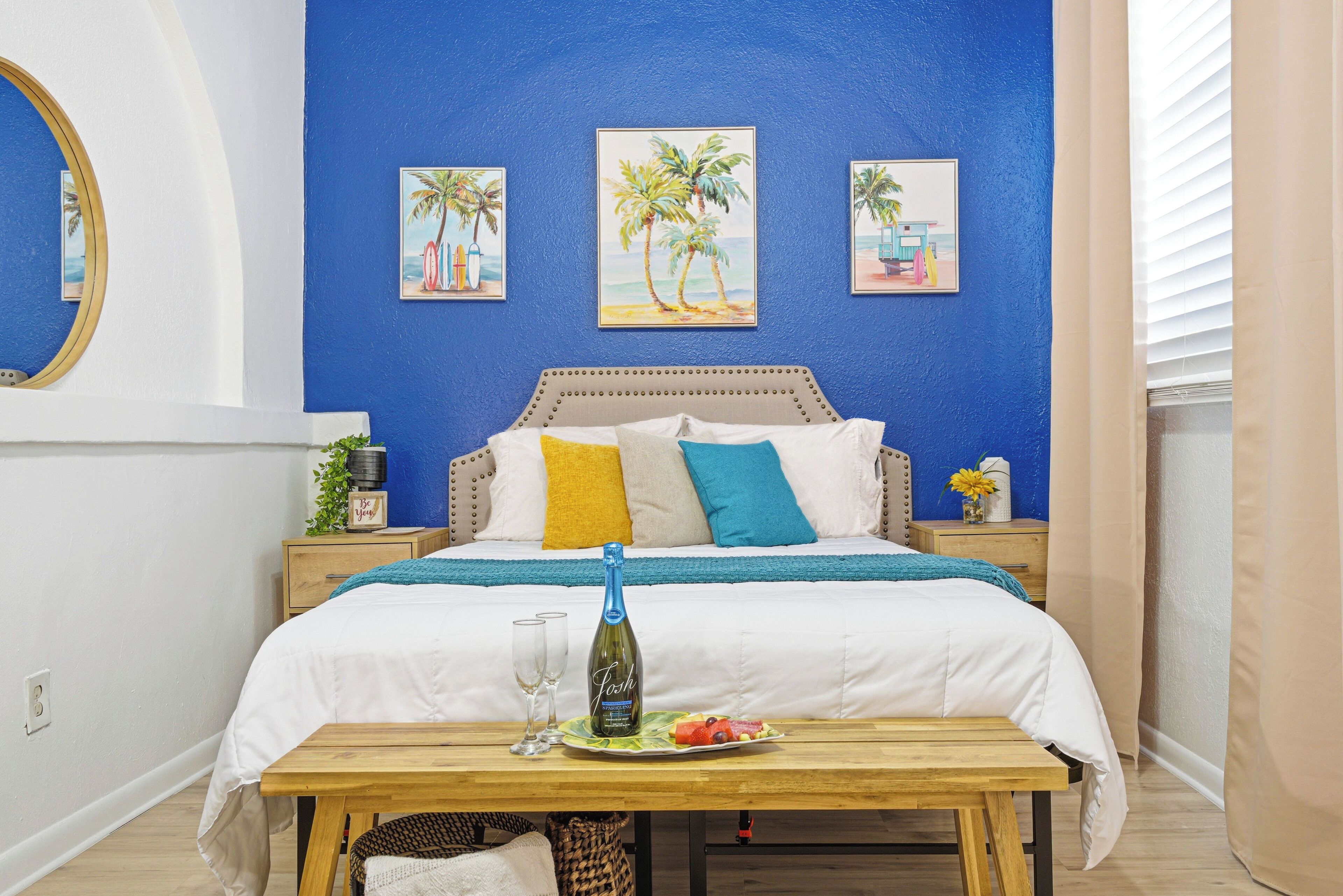 Un dormitorio con cama, mesitas de noche, una mesita auxiliar, una mesa de madera con una botella y vasos, y una pared decorada con tres cuadros de palmeras.