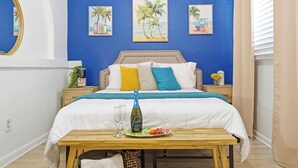 Ein Schlafzimmer mit einem Bett, Nachttischen, einem Beistelltisch, einem Holztisch mit einer Flasche und Gläsern sowie einer Wand mit drei Palmenbildern.
