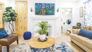 Ein Wohnzimmer mit Kamin, einem Stadtbildgemälde, einer großen Pflanze und einem blauen Teppich.