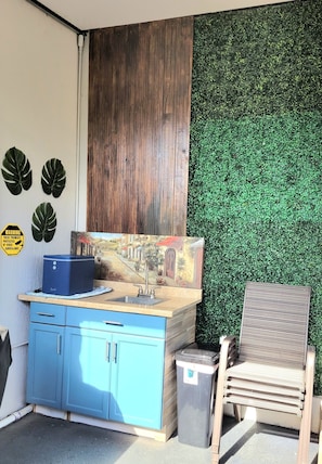 A cozy space with a cabinet, sink, and chair, featuring a framed picture and a trash bin.