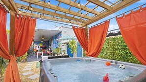 Ein Whirlpool unter einem hölzernen Pergola, umgeben von orangenen Vorhängen, mit einem Weg zu einem Gebäude mit farbenfrohen Wandgemälden.