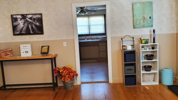 A room with a wooden floor, a welcome sign, a framed picture, a flower arrangement, a kitchen area visible through an open door, and a shelf with storage bins. - Make this house your HOME away from home (Meade)
