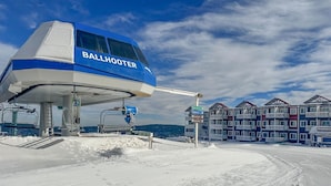 Estación de telesilla llamada BALLHOOTER, con suelo cubierto de nieve y un edificio de varios pisos al fondo.