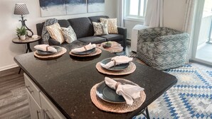 Una sala moderna con un comedor de color marrón oscuro para cuatro personas, un sofá gris con cojines estampados, un sillón con estampado y un cuadro abstracto grande en la pared.