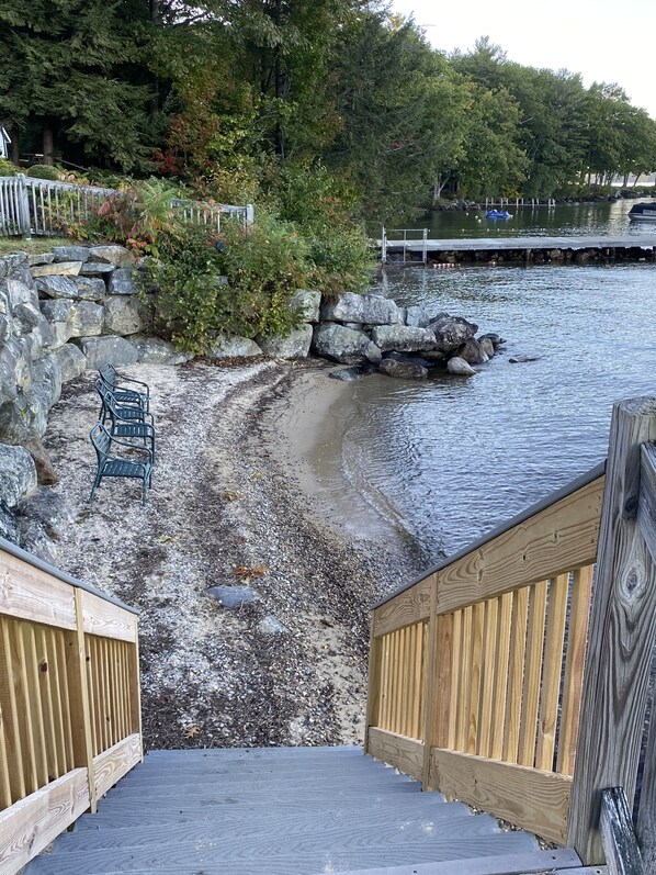 A wooden deck leading to a rocky shoreline with a chair and a dock extending into the water.