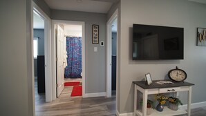Ein Flur mit einem 'Welcome'-Schild, einem an der Wand befestigten Fernseher, einem Konsolentisch mit einer Uhr und einem roten Teppich, der zu einem Raum mit einer gemusterten Duschvorhang führt.