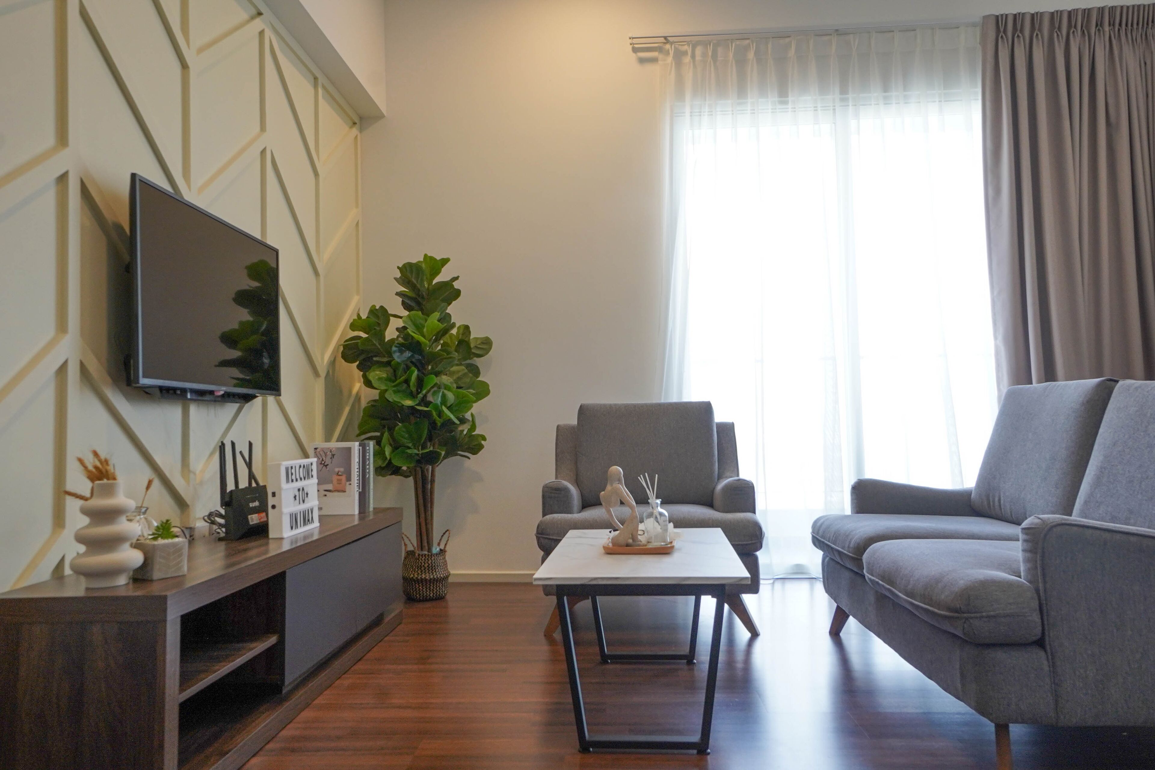 A modern living room with a flat-screen TV, a grey sofa, a coffee table, and a large plant.