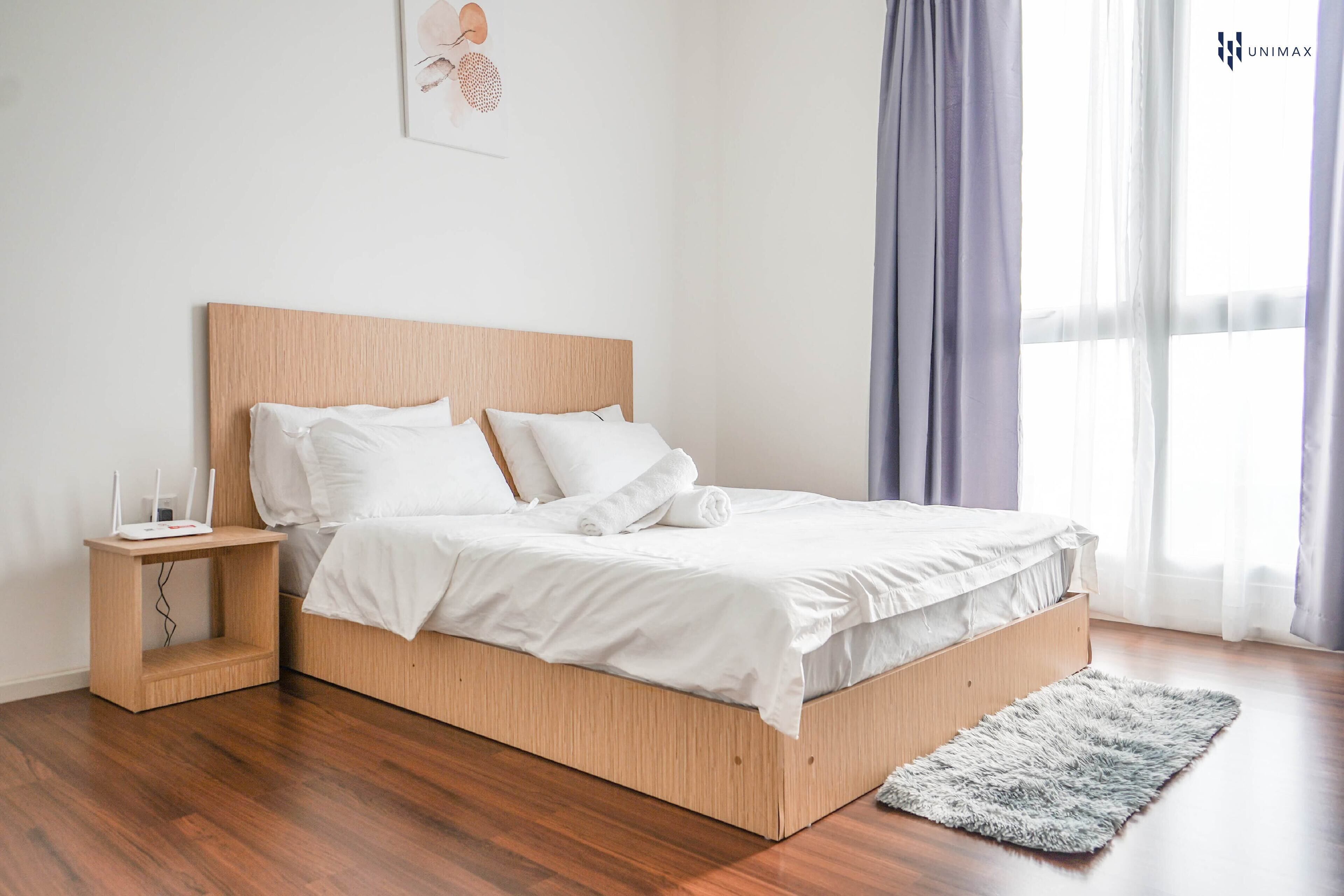 Een moderne slaapkamer met een houten bedframe, wit beddengoed, een nachtkastje met een lamp en een groot raam met gordijnen.