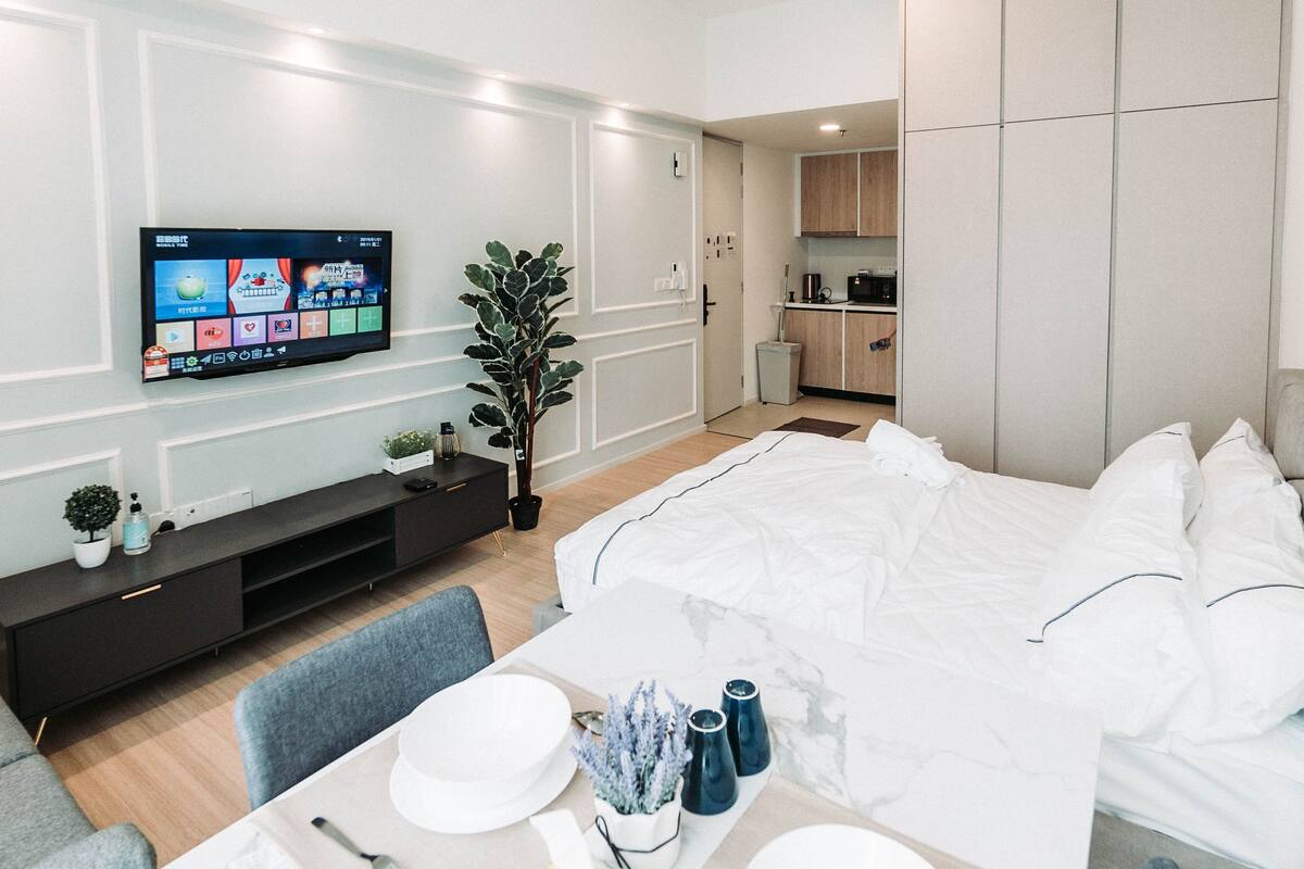 A modern hotel room with a bed, a TV, a small kitchenette, and a dining area.