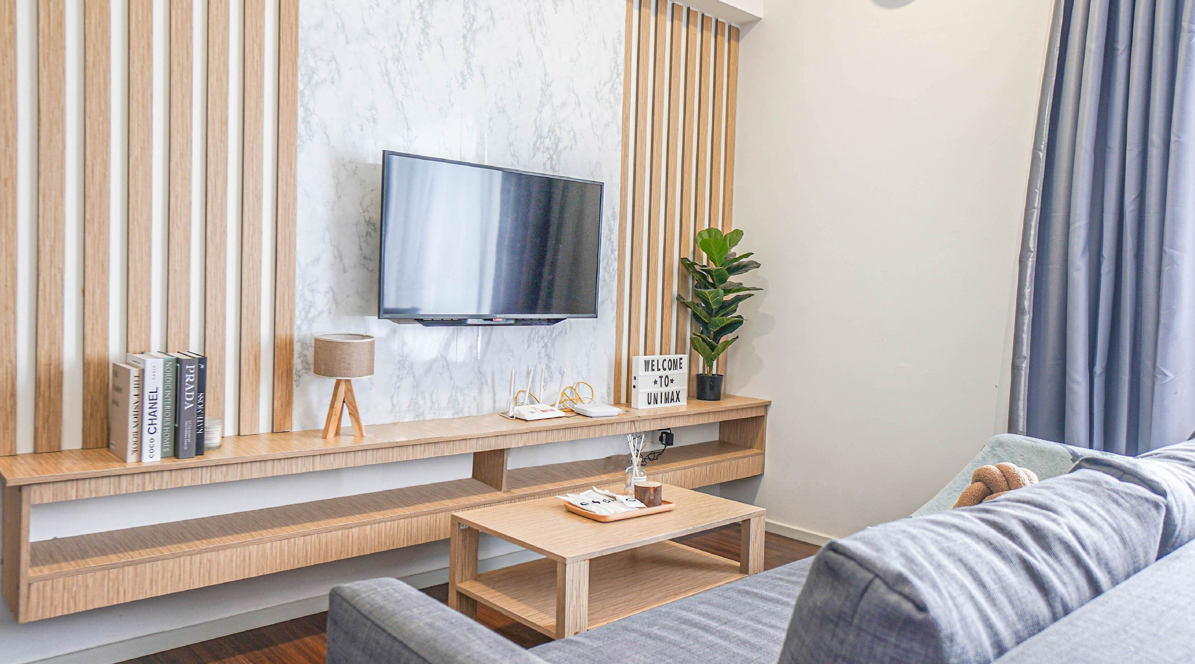 Ett modernt vardagsrum med en platt-TV som är monterad på en vägg av marmor, ett träbord med böcker och en lampa, ett soffbord och en grå soffa.
