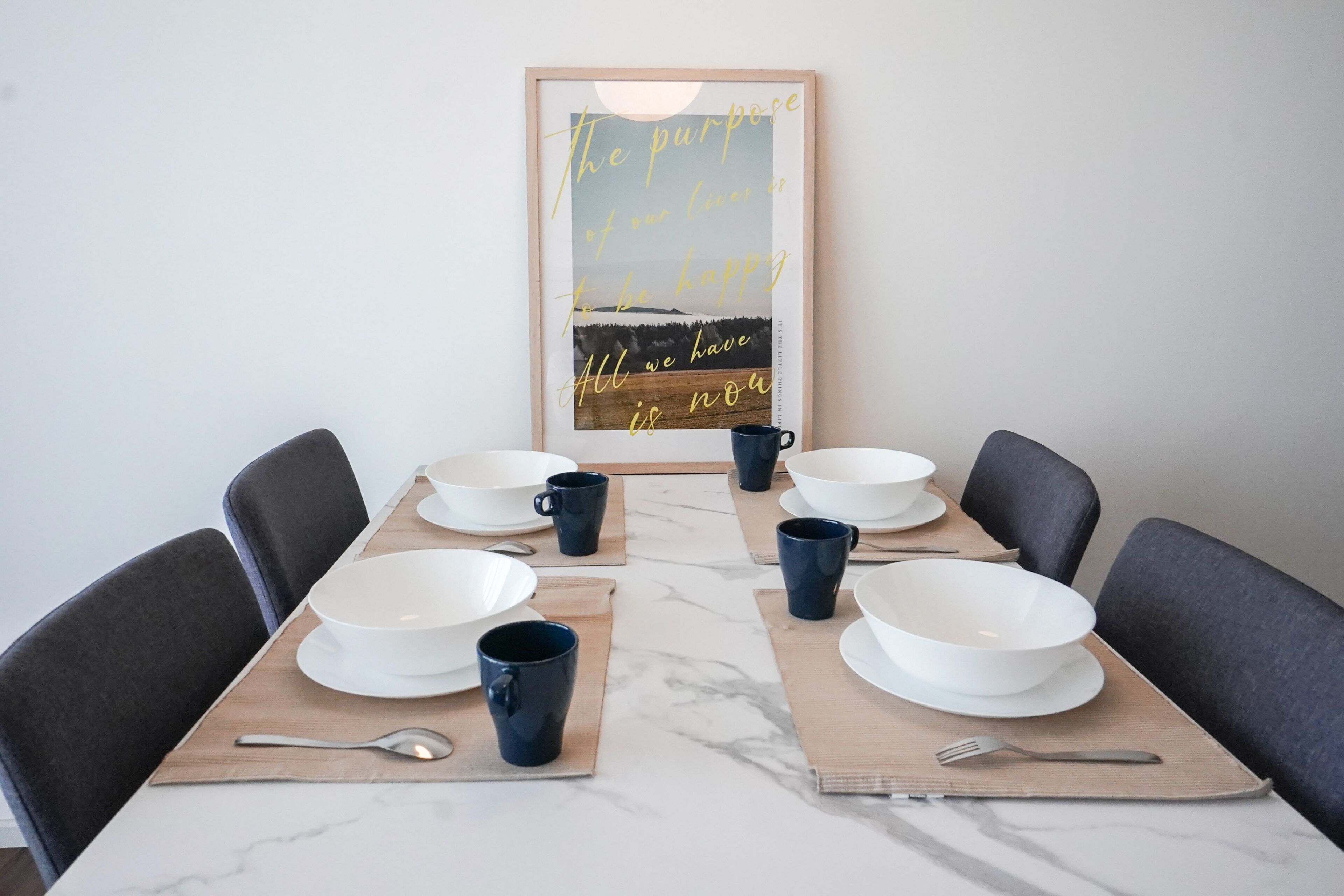 A dining table set with white bowls, blue mugs, and cutlery, with a framed inspirational quote on the wall behind.