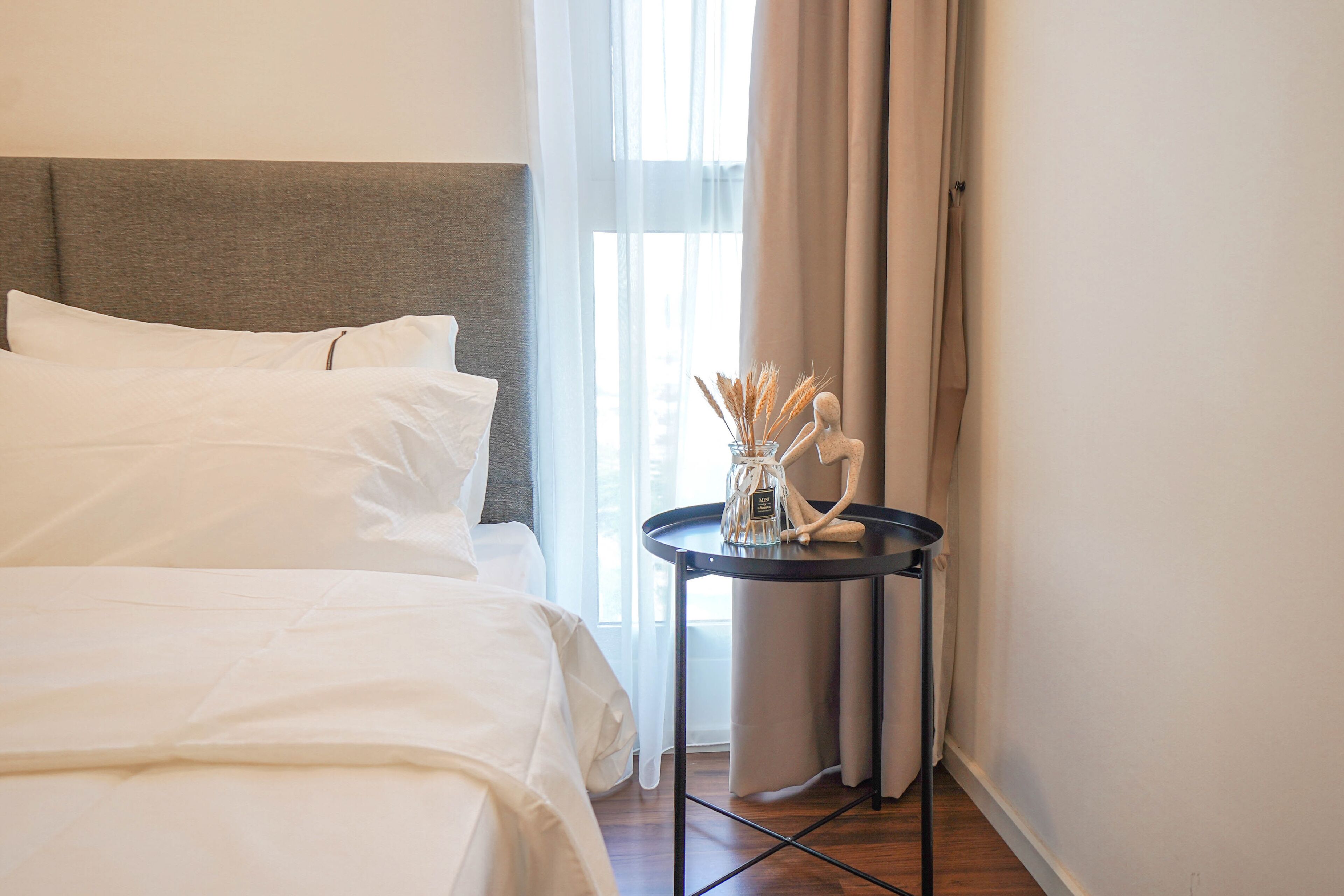 Una cama impecablemente hecha con sábanas blancas, una pequeña mesa redonda con un florero de plantas secas, y vistas a una ventana con cortinas traslúcidas.