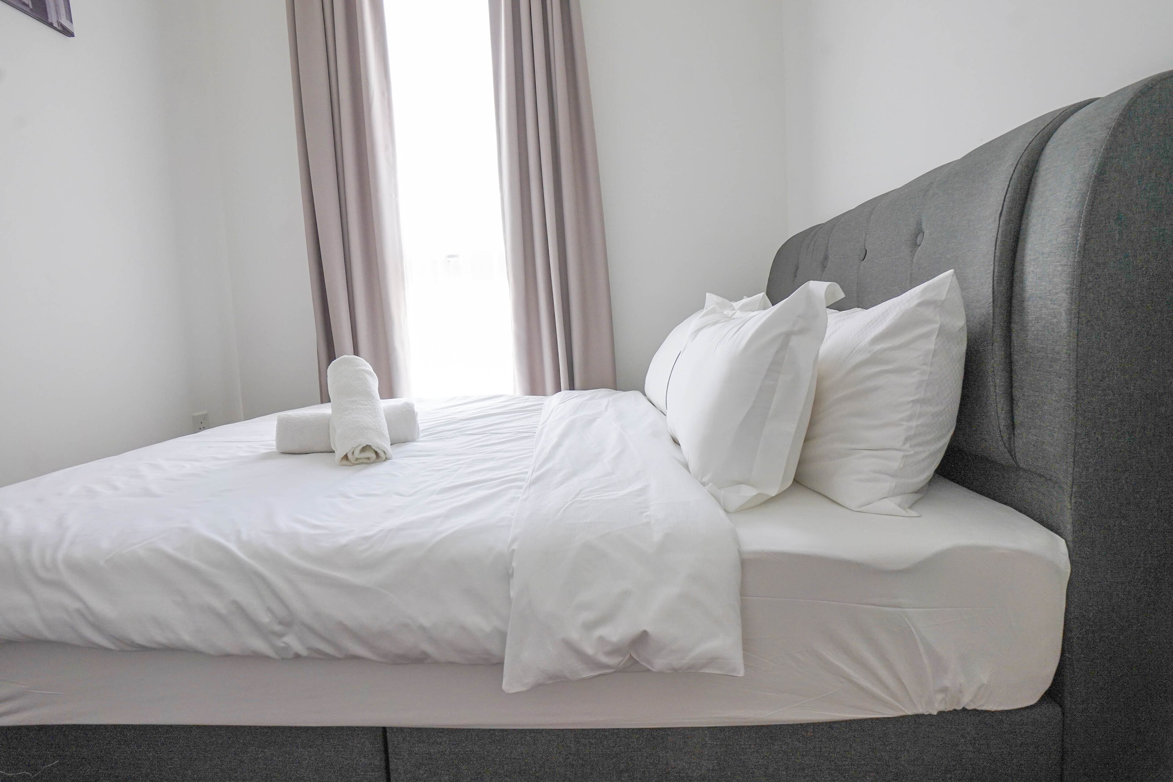 Una cama bien hecha con sábanas blancas y almohadas, un cabecero tapizado en gris, y una ventana con cortinas.