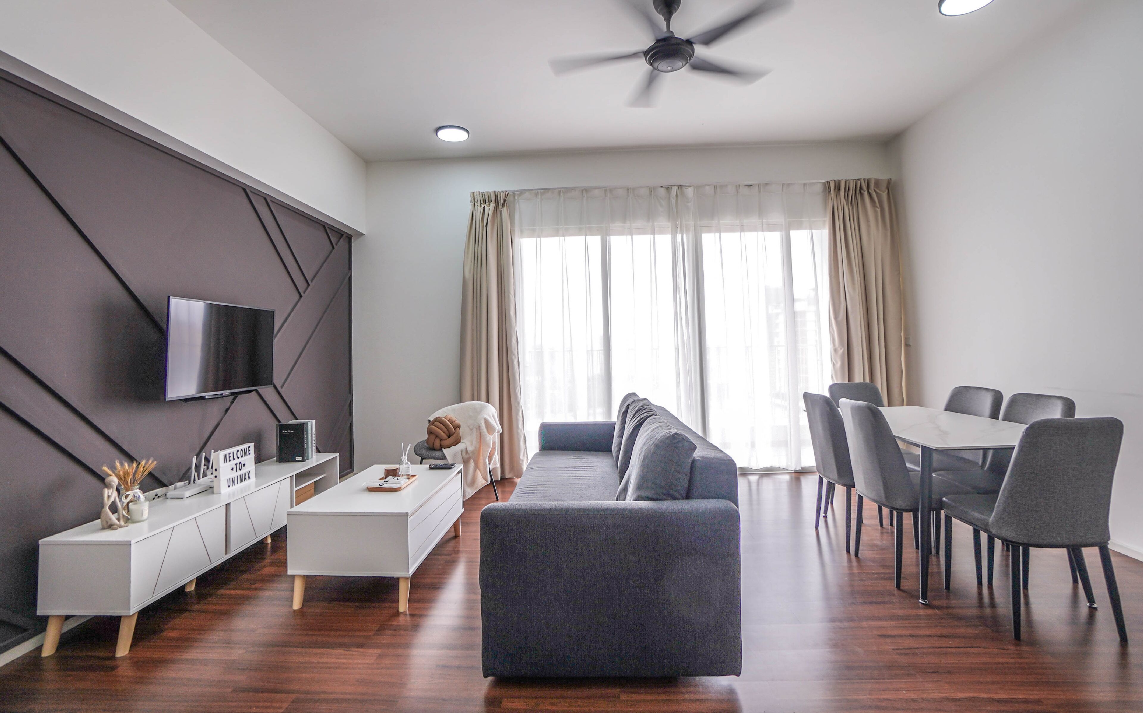 A modern living room with a sofa, dining table, and chairs, a TV mounted on the wall, and a ceiling fan.