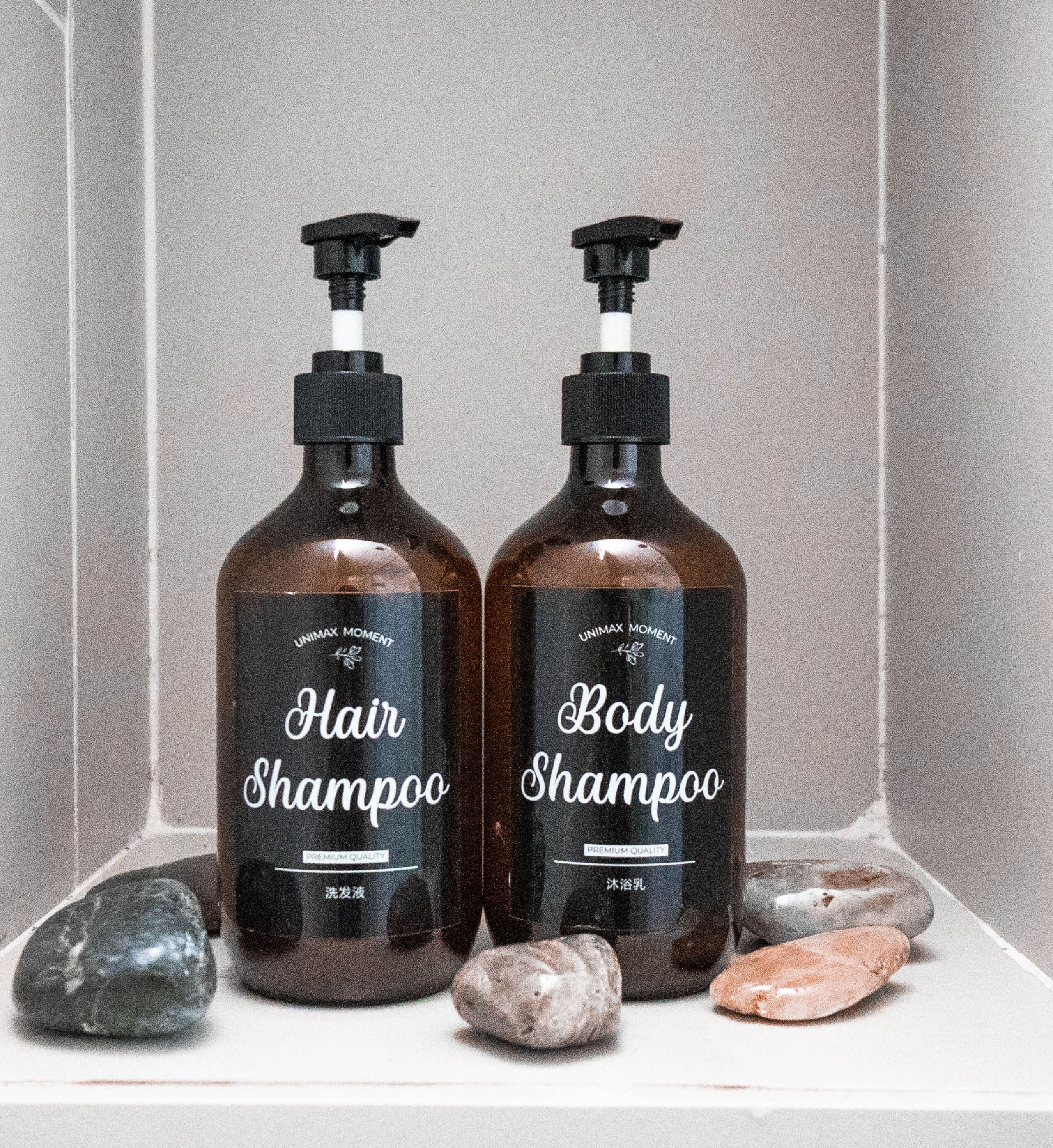 Two bottles of hair and body shampoo with pumps, placed on a shelf with stones.