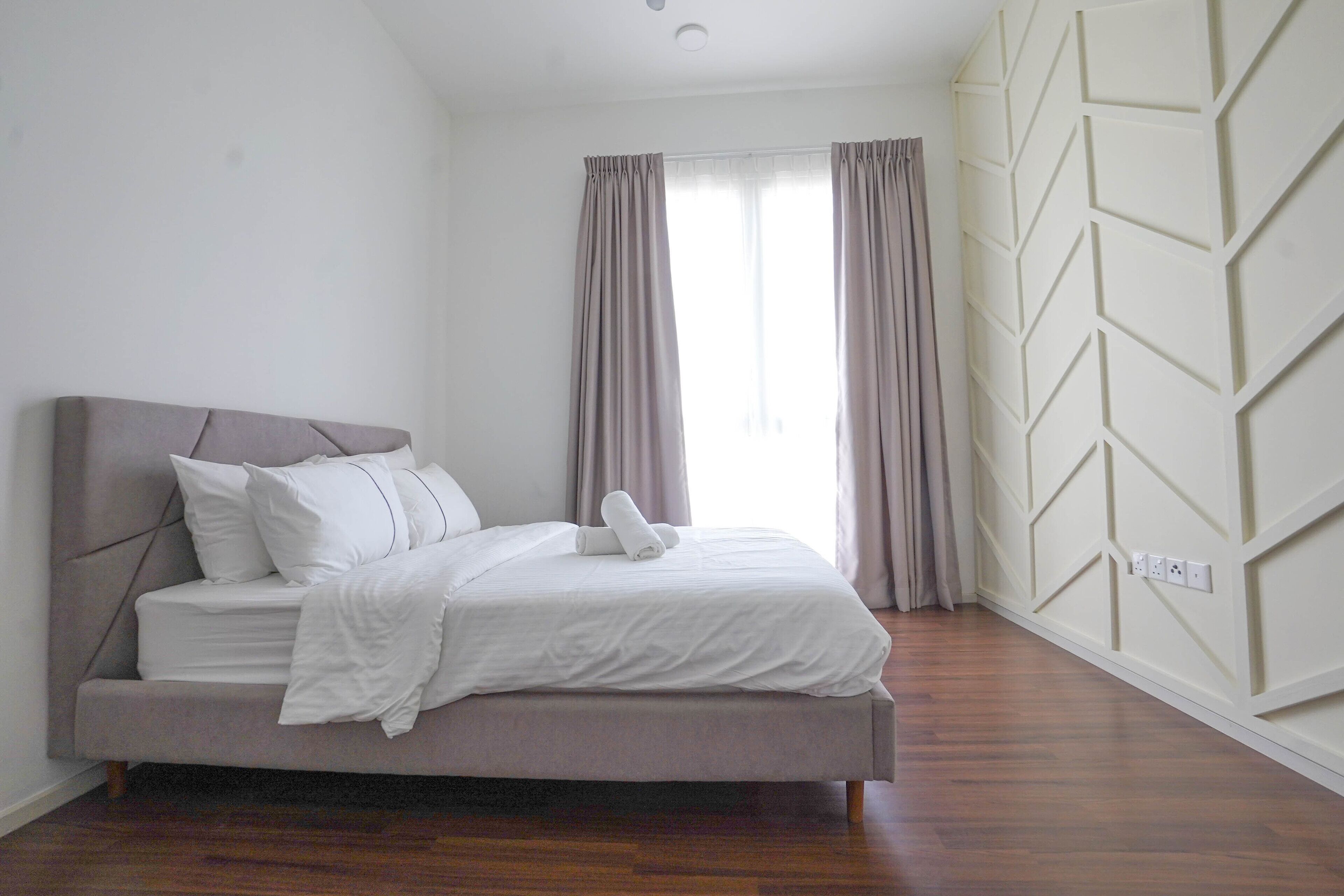 A neatly made bed with a headboard, a window with curtains, and a wall with a geometric pattern.