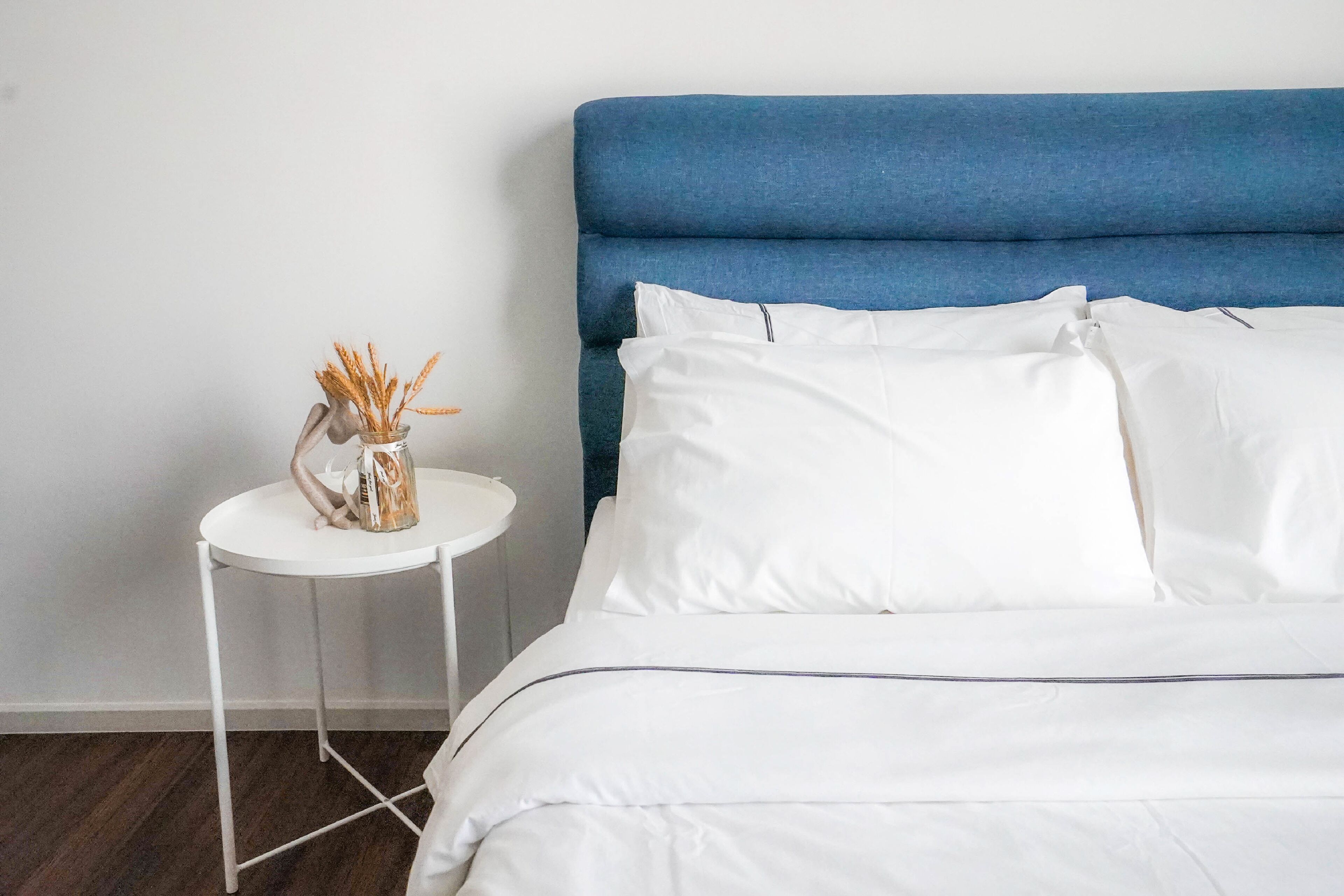 Een keurig opgemaakt bed met witte lakens en een blauw hoofdeinde, een klein wit nachtkastje met een vaas vol korellen, en een effen witte muur.
