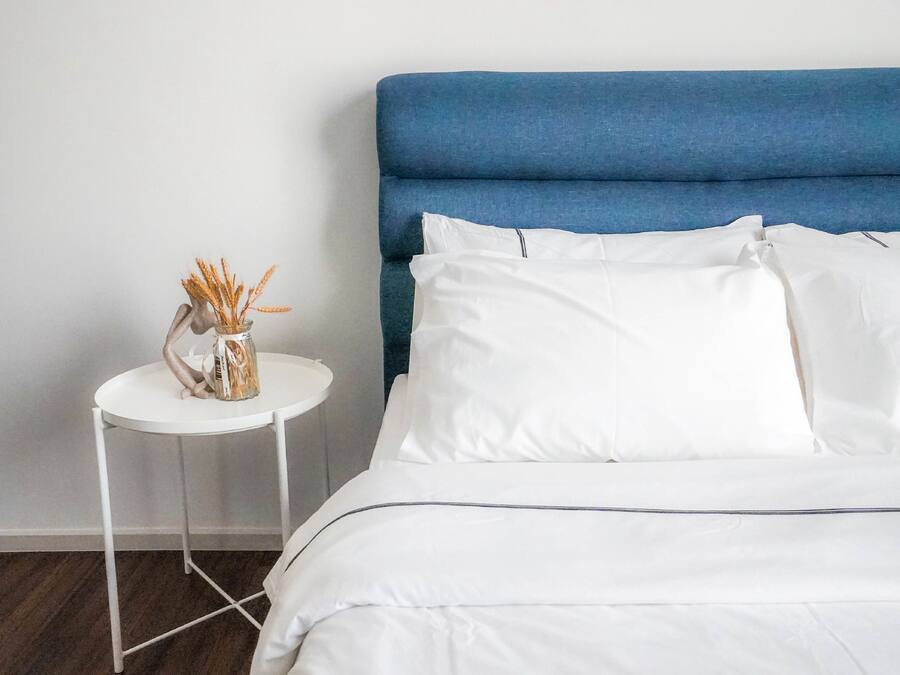 Een keurig opgemaakt bed met witte lakens en een blauw hoofdeinde, een klein wit nachtkastje met een vaas vol korellen, en een effen witte muur.