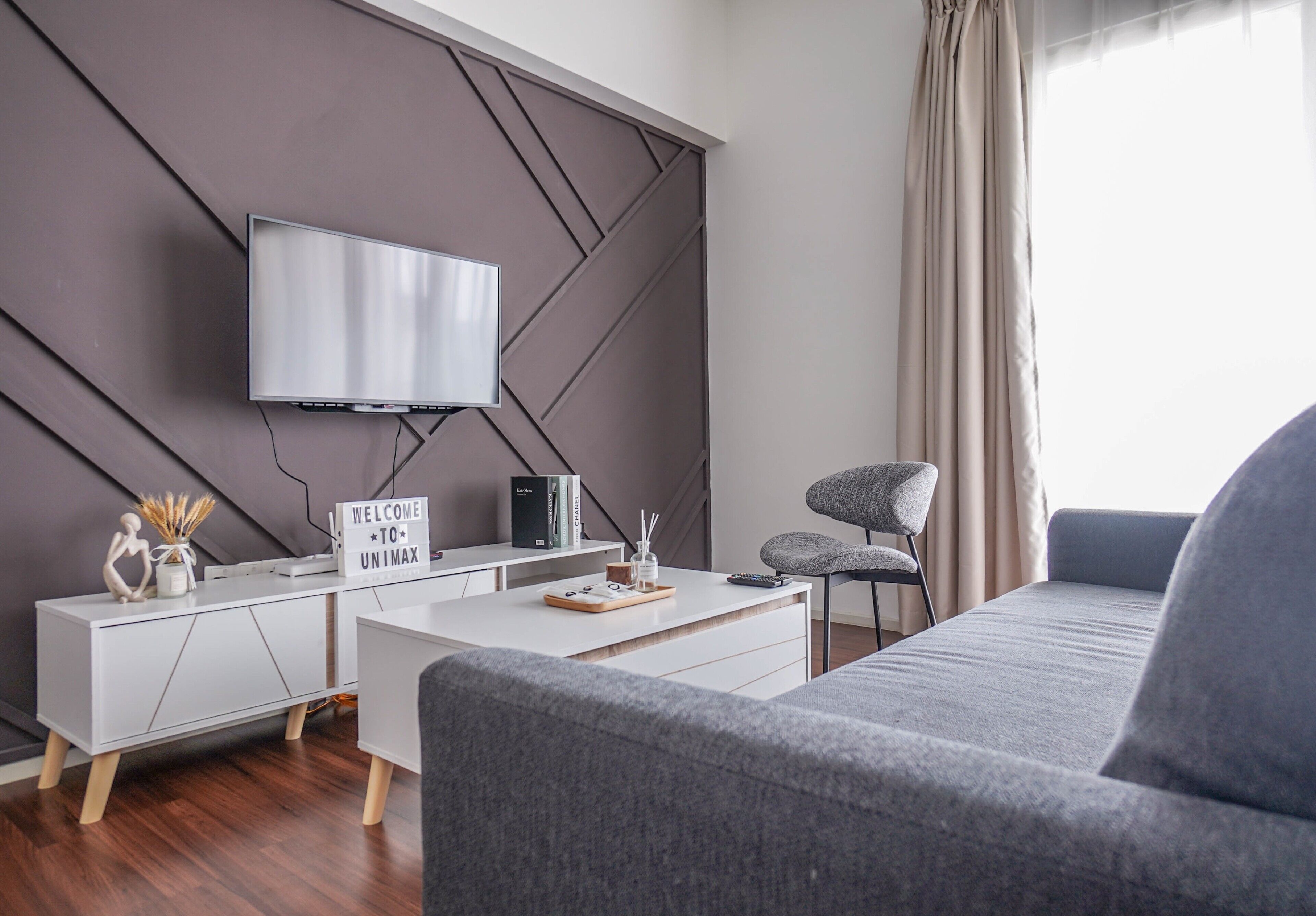 Una sala de estar moderna con un televisor de pantalla plana montado en una pared con diseño geométrico, una mesita blanca con una bandeja, un sofá gris y un sillón gris.